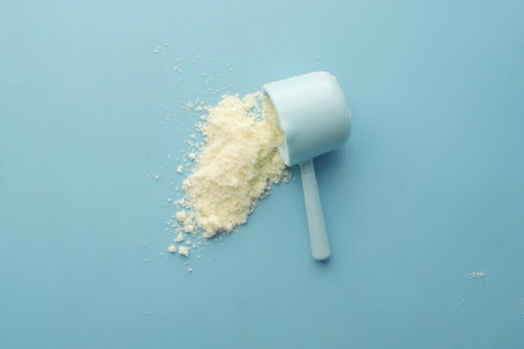 White scooper laying on its side, with white supplement powder spilled out, on a light blue background.