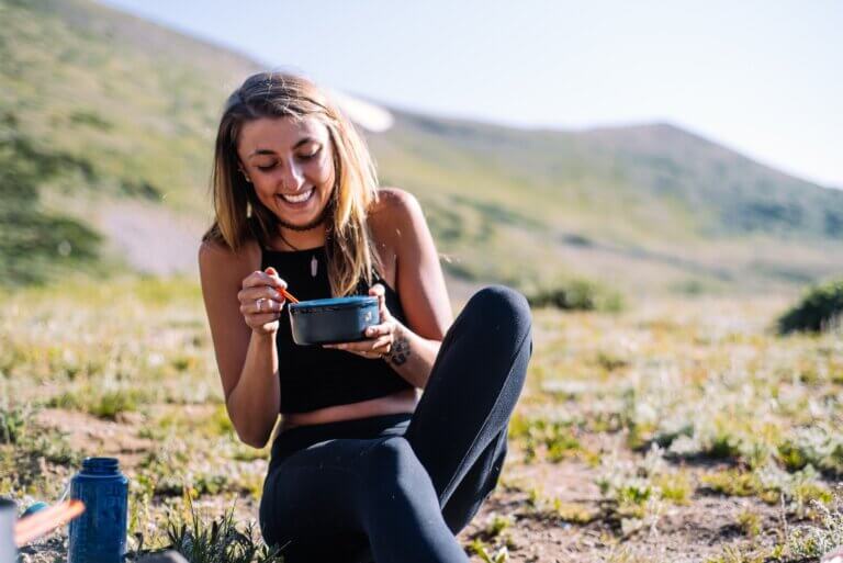 woman in black tank top and black pants sitting on ground with mountains in the background, smiling while eating from a blue bowl