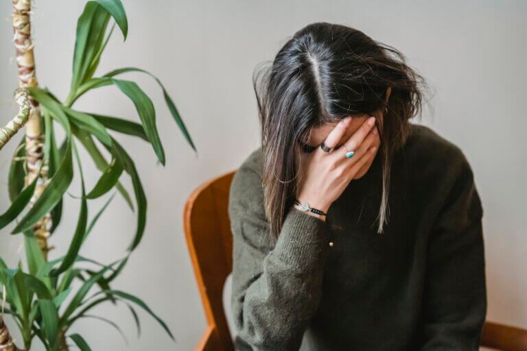 Young upset woman with dark hair wearing sweater covering face with hand while sitting on chair next to a houseplant
