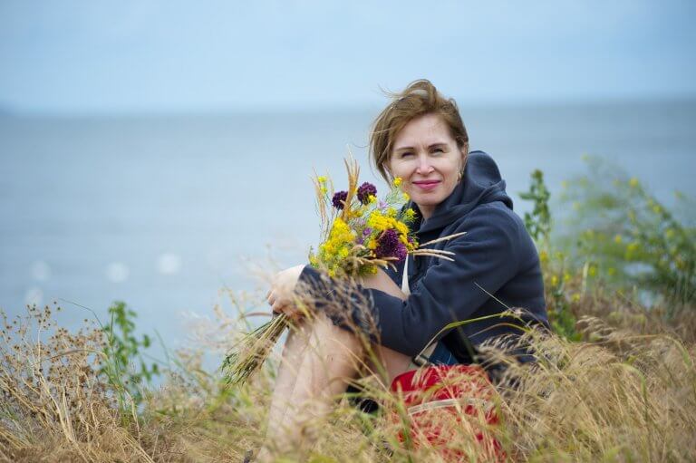 Woman Sitting on Grass Holding Flowers Wearing Black Hoodie With Water In Background