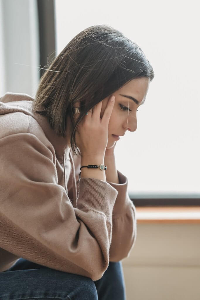 Side view of sad woman sitting down wearing a light brown sweatshirt holding her head and looking down