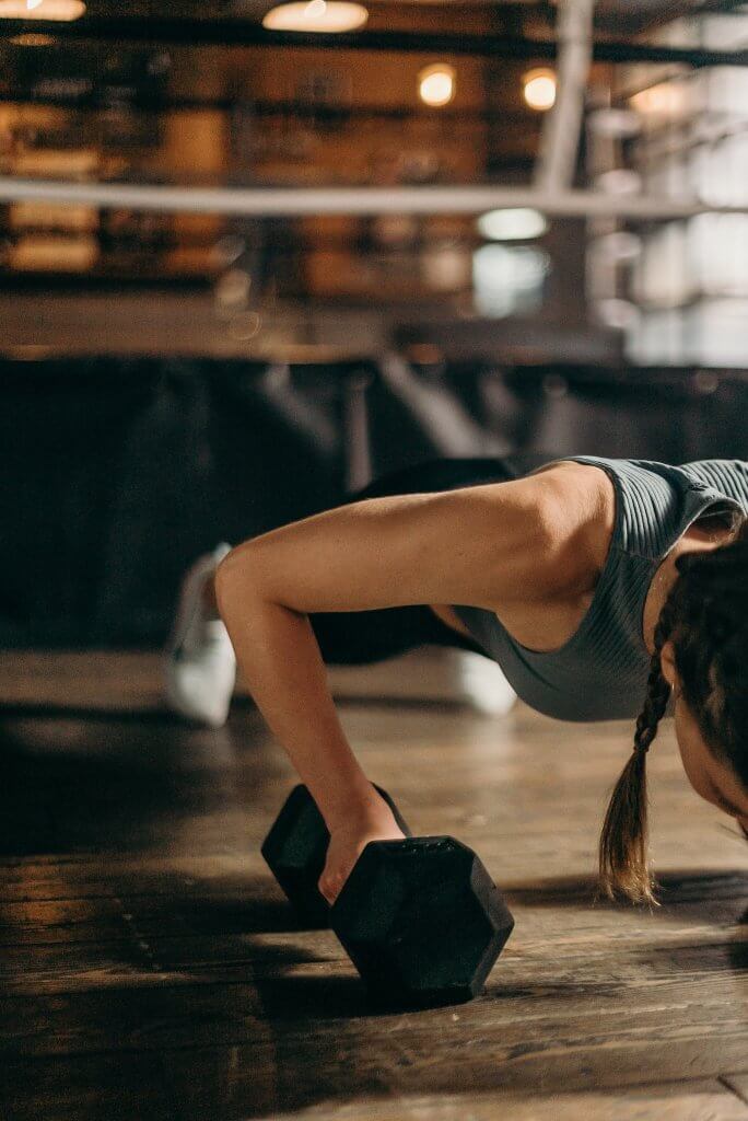 Woman in Black and White Tank Top on Brown Floor in Pushup Position With Hands on  Black Dumbbells