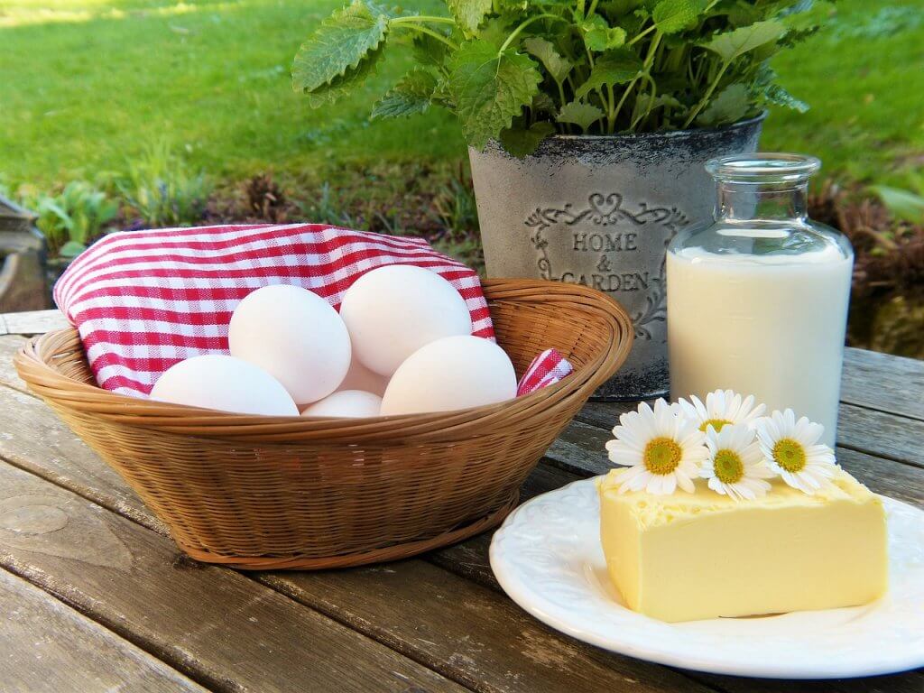 Brown basket filled with eggs on a red and white checkered cloth, a glass jar of milk, and a piece of cheese on a white plate with daisies on top, sitting on top of a wooden table surface