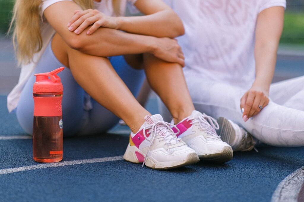 Anonymous view of two women in sports attire sitting down on the ground after training, with orange water bottle